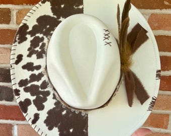 May include: A two-tone fedora hat with a cowhide pattern on one side and a solid white color on the other. The hat features brown feathers and a brown ribbon. The brim is stitched with white thread. The text "XXXX" is embroidered on the crown.