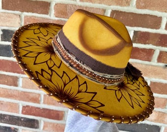 May include: A yellow cowboy hat with a brown brim and a sunflower design. The hat features a brown band with decorative stitching and a tassel. The brim is decorated with white beads and brown stitching.