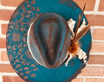May include: A teal blue cowboy hat with brown accents and a floral design. The hat has a brown leather band with a white lace trim and a brown feather accent.