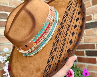 May include: A tan felt hat featuring a wide brim with a dark brown geometric diamond and zigzag pattern burned into the surface. The crown is wrapped with a layered band of frayed fabric in turquoise, red, and white, accented by a gold chain.