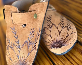 May include: A pair of tan suede slip-on clogs featuring dark brown floral line art on the toe area. Each shoe has an adjustable strap with a brass-colored metal rivet and a cork-textured footbed with the partial text FASHION visible.