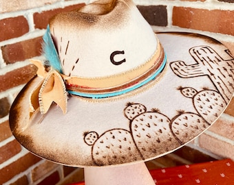 Puede incluir: Un sombrero de vaquero blanco con un ala marrón y un diseño quemado. El sombrero tiene una banda de cuero marrón con un ribete turquesa y una banda beige con una raya roja. El sombrero está decorado con un diseño de cactus y la letra "C" está bordada en la corona.
