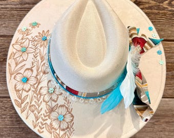 May include: Cream-colored fedora hat with floral and turquoise accents. The hat features a painted floral design in brown, turquoise embellishments, and a patterned band. A blue feather and patterned fabric add a decorative touch.