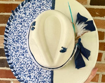 May include: Cream-colored fedora hat with a wide brim. The brim features a section with a blue floral pattern and a denim trim. The hat is decorated with a feather, a blue bow, and a small blue floral accent.