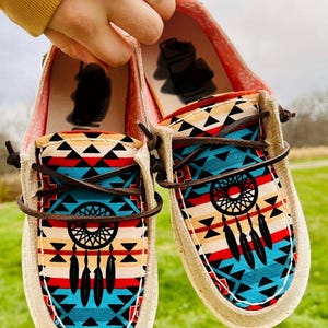 May include: A pair of canvas slip-on shoes with a Native American inspired pattern featuring a dreamcatcher design in red, blue, white, and black. The shoes have brown laces.