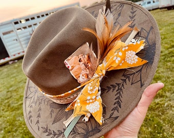 Puede incluir: Un sombrero de fieltro marrón con ala ancha, decorado con un lazo de tela floral amarillo, una pluma y una foto vintage. El sombrero tiene un diseño floral negro en el ala.