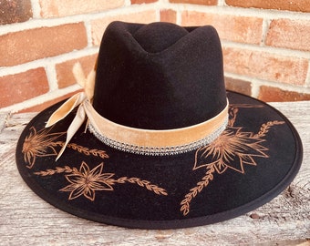 Puede incluir: Un sombrero fedora de fieltro negro con ala ancha. El sombrero tiene una banda de terciopelo marrón con una fila de diamantes de imitación y un diseño floral marrón en el ala.