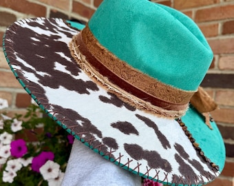May include: A teal felt fedora hat featuring a wide brim with a brown and white cow print pattern. The hat is accented with a brown paisley patterned ribbon, a frayed fabric trim, and a decorative row of small dark brown beads.