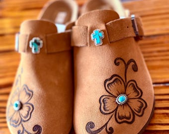 May include: A pair of tan suede clogs featuring a dark brown floral line art design on the toe. Each shoe is accented with a small turquoise stone in the center of the flower and a turquoise cross-shaped charm attached to the adjustable strap.
