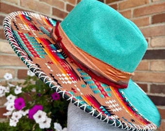 May include: A wide-brimmed fedora hat featuring a vibrant turquoise crown and a tan brim adorned with a colorful geometric pattern. The hat includes a layered band in shades of orange and red, with white whipstitching along the outer edge.