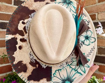 May include: A beige fedora hat featuring a split-design brim with a brown and white cow print on one side and teal floral line art on the other. The hat is accented with a brown ribbon, teal feathers, and a white lace trim around the crown.