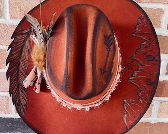 May include: A rust-colored cowboy hat with a decorative feather and fabric arrangement on the side. The hat features an arrow design and mountain range illustration. The brim is trimmed with white lace.