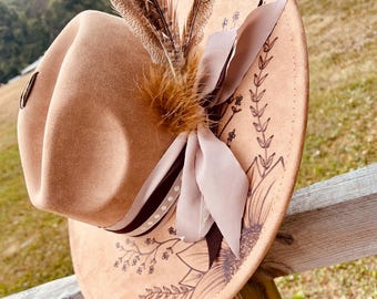 May include: A tan fedora hat with a decorative feather and ribbon arrangement. The hat features a floral design around the brim. The hat is resting on a wooden fence.