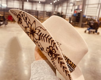May include: A beige cowboy hat with a floral design and a leopard print band. The hat is decorated with rhinestones.