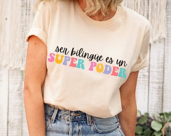 Op de afbeelding: Een beige T-shirt met de tekst "ser bilingüe es un SUPER PODER" in regenboogkleuren. De tekst is in een retro-lettertype stijl.