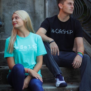 Puede incluir: Dos personas est&aacute;n sentadas en un conjunto de escalones de concreto. La persona de la izquierda lleva una camiseta turquesa con el texto "Be Salt" y "Matthew 5:13" impreso en ella. La persona de la derecha lleva una camiseta negra con el texto "WORSHIP" impreso en ella.