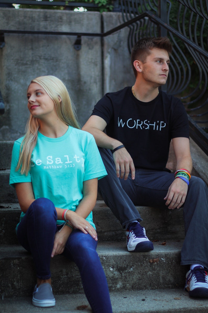 Puede incluir: Dos personas est&aacute;n sentadas en un conjunto de escalones de concreto. La persona de la izquierda lleva una camiseta turquesa con el texto "Be Salt" y "Matthew 5:13" impreso en ella. La persona de la derecha lleva una camiseta negra con el texto "WORSHIP" impreso en ella.