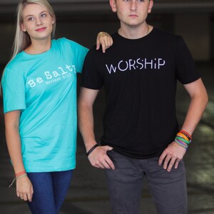 Puede incluir: Dos personas vistiendo camisetas con mensajes religiosos. La persona de la izquierda lleva una camiseta turquesa con el texto "Be Salty Matthew 5:13". La persona de la derecha lleva una camiseta negra con el texto "WORSHIP".