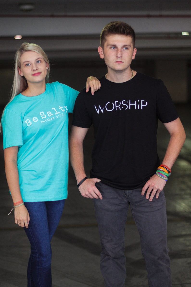 Puede incluir: Dos personas vistiendo camisetas con mensajes religiosos. La persona de la izquierda lleva una camiseta turquesa con el texto "Be Salty Matthew 5:13". La persona de la derecha lleva una camiseta negra con el texto "WORSHIP".