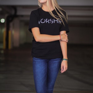 Puede incluir: Una mujer con una camiseta negra con la palabra "WORSHIP" impresa en blanco en la parte delantera. Lleva pantalones vaqueros azules y tiene el pelo rubio.