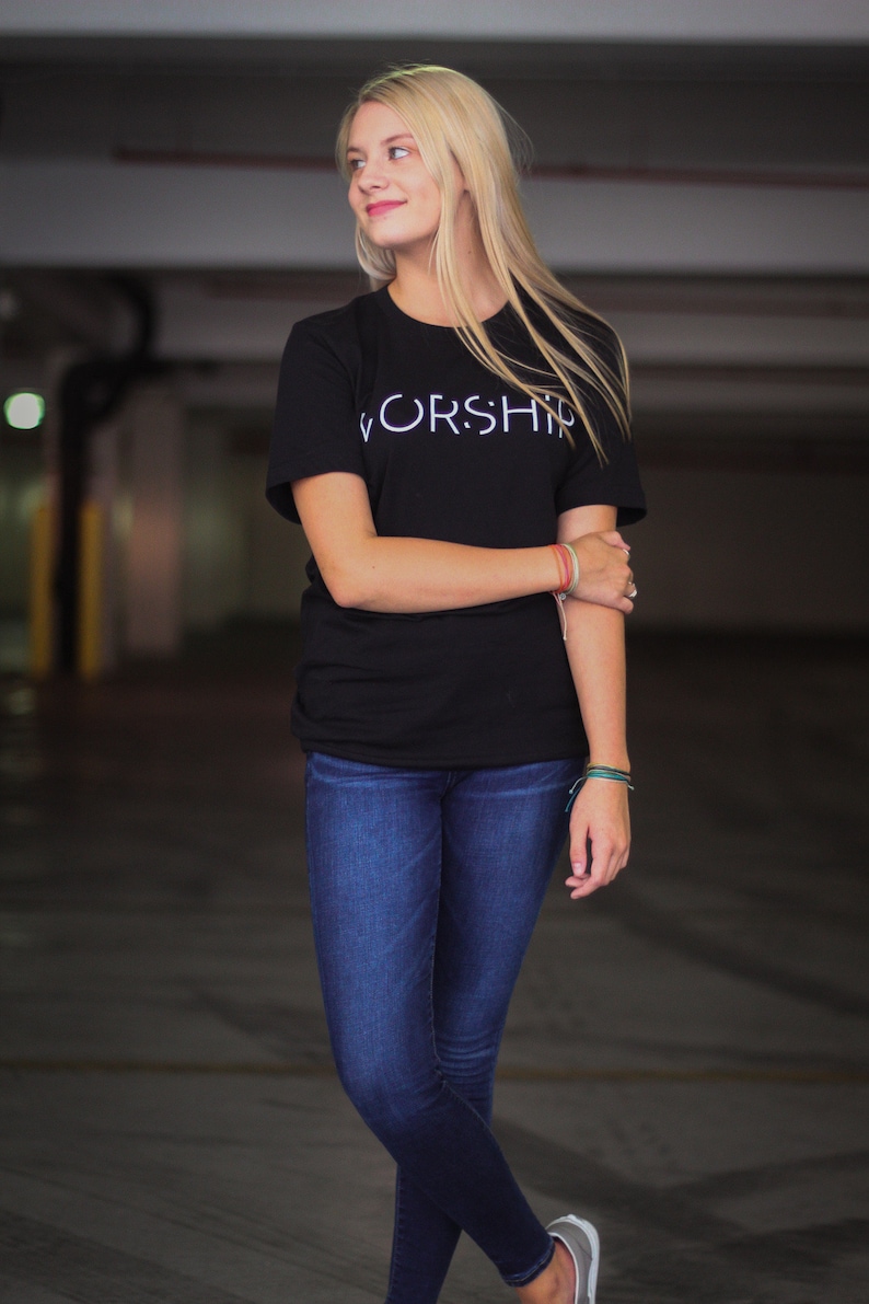 Puede incluir: Una mujer con una camiseta negra con la palabra "WORSHIP" impresa en blanco en la parte delantera. Lleva pantalones vaqueros azules y tiene el pelo rubio.