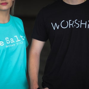 Puede incluir: Dos personas vistiendo camisetas con mensajes religiosos. La persona de la izquierda lleva una camiseta turquesa con el texto "Be Salt" y "Matthew 5:13". La persona de la derecha lleva una camiseta negra con el texto "WORSHIP" en blanco.