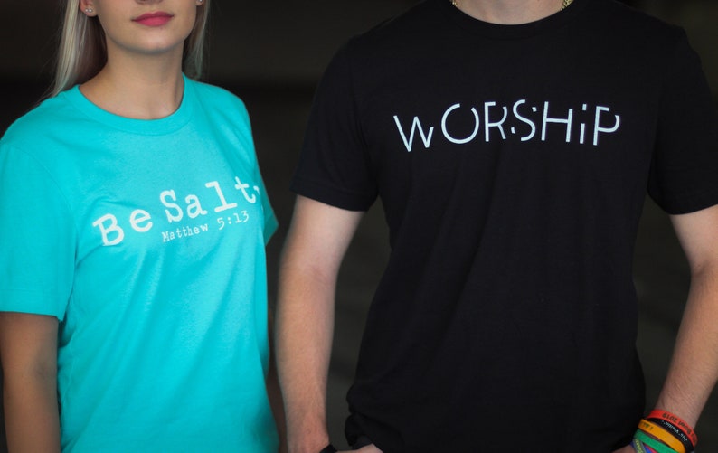 Puede incluir: Dos personas vistiendo camisetas con mensajes religiosos. La persona de la izquierda lleva una camiseta turquesa con el texto "Be Salt" y "Matthew 5:13". La persona de la derecha lleva una camiseta negra con el texto "WORSHIP" en blanco.
