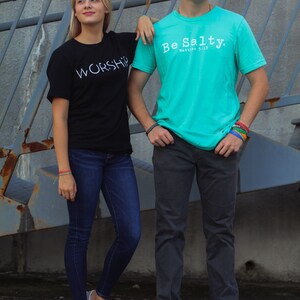 Puede incluir: Dos personas vistiendo camisetas con texto. La persona de la izquierda lleva una camiseta negra con la palabra "WORSHIP" en texto blanco. La persona de la derecha lleva una camiseta turquesa con el texto "Be Salty. Matthew 5:13".