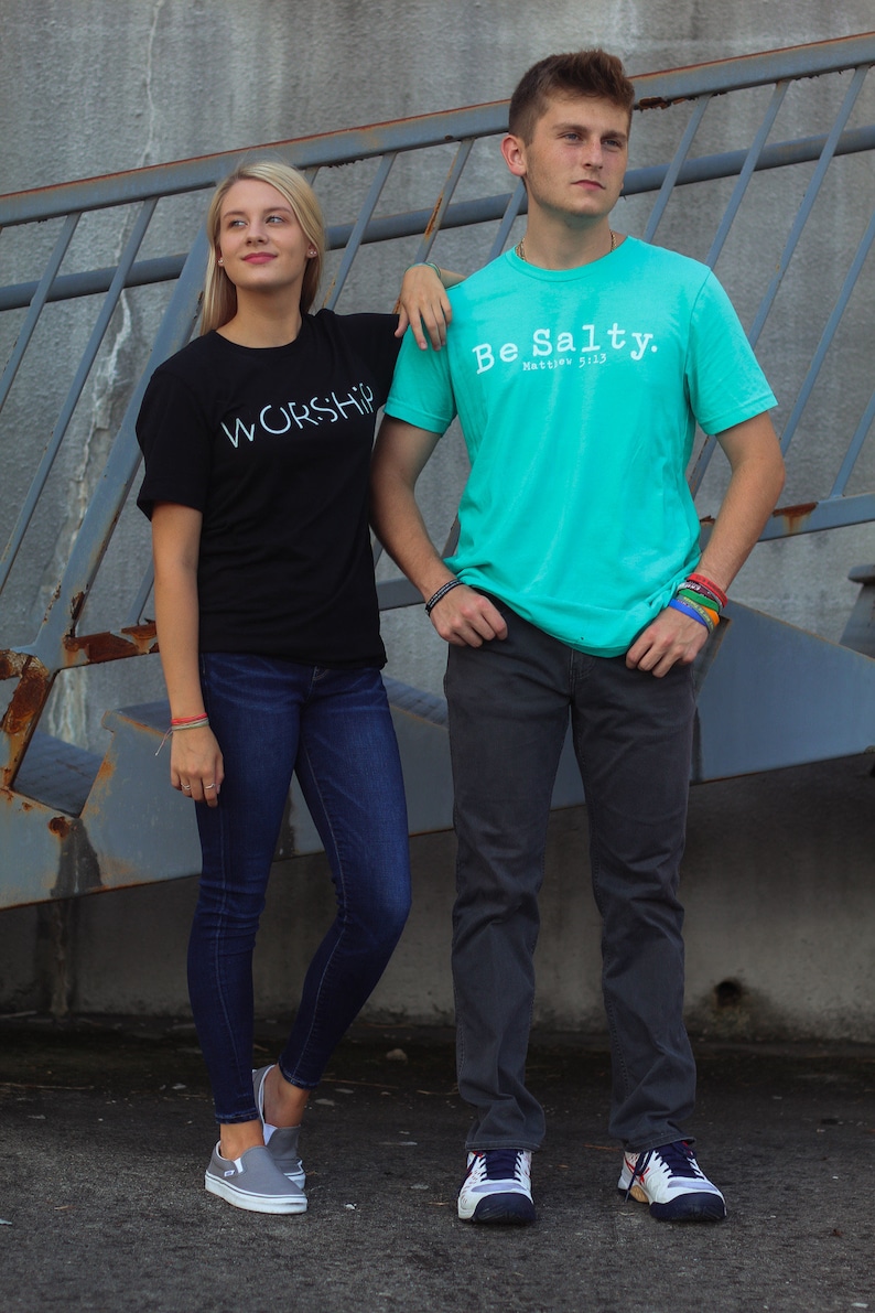 Puede incluir: Dos personas vistiendo camisetas con texto. La persona de la izquierda lleva una camiseta negra con la palabra "WORSHIP" en texto blanco. La persona de la derecha lleva una camiseta turquesa con el texto "Be Salty. Matthew 5:13".