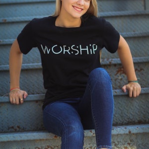 Puede incluir: Una mujer con una camiseta negra con la palabra "WORSHIP" impresa en blanco en la parte delantera. Est&aacute; sentada en un tramo de escaleras de metal y lleva pantalones vaqueros azules.