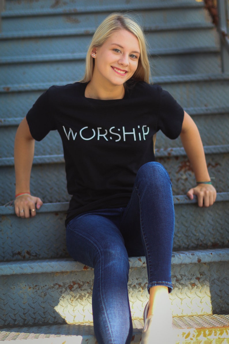 Puede incluir: Una mujer con una camiseta negra con la palabra "WORSHIP" impresa en blanco en la parte delantera. Est&aacute; sentada en un tramo de escaleras de metal y lleva pantalones vaqueros azules.