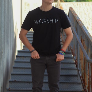 Puede incluir: Una persona joven lleva una camiseta negra con la palabra "WORSHIP" impresa en blanco en la parte delantera. Est&aacute; de pie en un tramo de escaleras de metal.