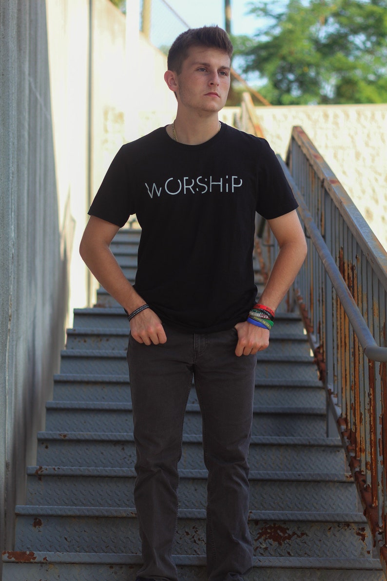 Puede incluir: Una persona joven lleva una camiseta negra con la palabra "WORSHIP" impresa en blanco en la parte delantera. Est&aacute; de pie en un tramo de escaleras de metal.