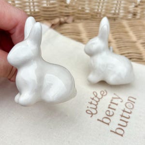 May include: Two white ceramic rabbit-shaped drawer knobs with a glossy finish. One knob is held in the foreground, while the other sits on a light-coloured fabric pouch printed with the text little berry button in a brown script font.