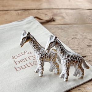 May include: Two white ceramic giraffe figurines with gold accents. The giraffes are standing on a white canvas bag with the text "little berry button" printed on it.