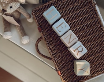 May include: A brown woven wicker suitcase with a handle, topped with five light-colored wooden blocks arranged to spell the name HENRY in gold lettering. Beside the suitcase sits a small, light brown crocheted stuffed deer toy with white accents.