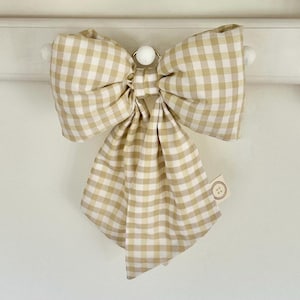 May include: A decorative bow featuring a beige and white gingham check pattern. The bow has two large loops and two long, hanging tails. A small, circular tag with a four-hole button graphic is attached to the right side of the fabric bow.