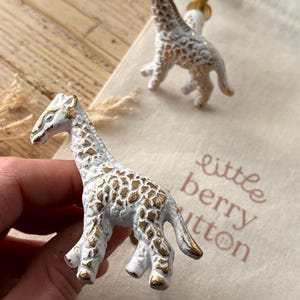 May include: Two white and gold giraffe drawer pulls with a gold screw. The drawer pulls are on a white fabric bag with the text "little berry button" printed on it.