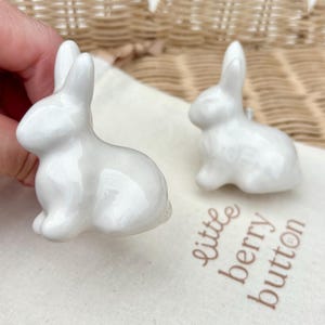 May include: Two white ceramic rabbit-shaped knobs with a glossy finish. One rabbit is held in the foreground, while the other sits on a cream-colored fabric pouch printed with the text little berry button in brown script lettering.