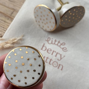 May include: Three white ceramic knobs with gold polka dots and a gold rim. The knobs have a screw on the back for attaching to a surface. The knobs are on a white fabric surface with the text "little berry button" printed on it.