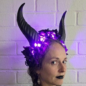 May include: A headpiece featuring two large, curved black horns and dark floral elements illuminated by bright purple LED lights. The structure is designed to be worn on the head, showcasing a contrast between the dark textures and the vibrant glow.