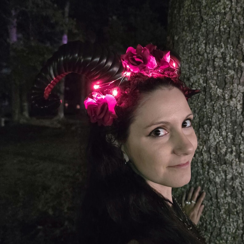 May include: A headband featuring large, curved black ram horns and dark pink artificial roses. The accessory is illuminated by small, bright pink LED fairy lights woven throughout the floral arrangement and around the base of the horns.