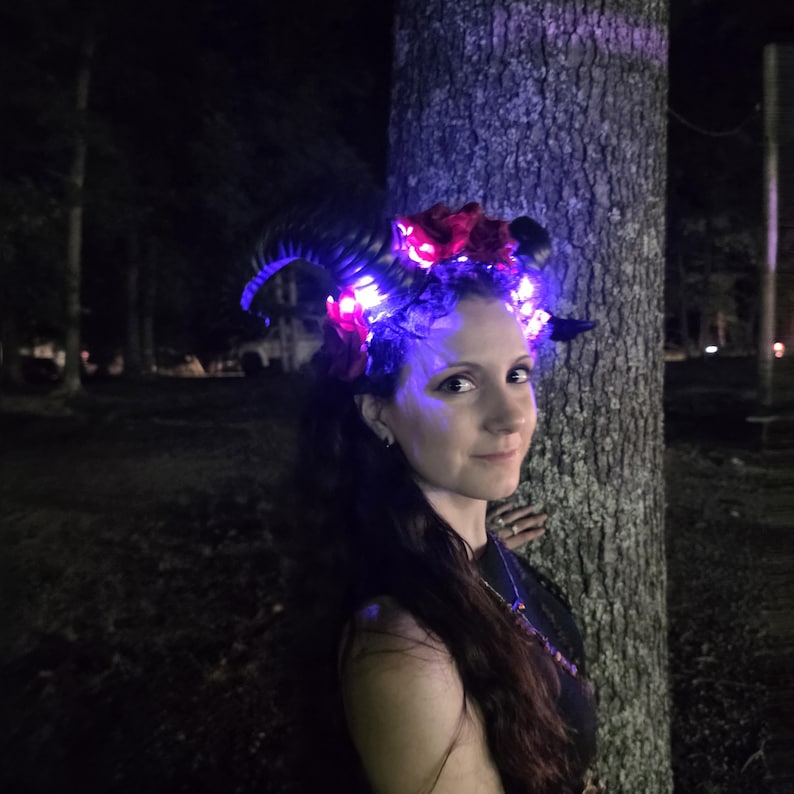 May include: A headpiece featuring two large, curved black horns adorned with dark red artificial roses and integrated bright purple and white LED lights that illuminate the floral elements and the surrounding area in a dark outdoor setting.