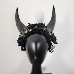 May include: Black headband with two large horns and a black floral garland with small lights. The headband is on a black mannequin head.