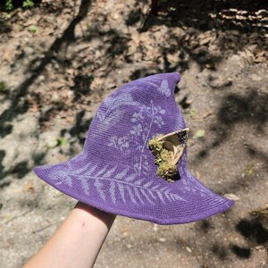 May include: A purple witch hat with a wide brim and a pointed crown. The hat features a floral pattern and is adorned with a small, decorative piece of wood and moss. The hat is made of a knitted fabric.