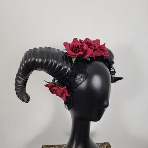 May include: A headpiece featuring large, curved black ram horns attached to a black lace headband. The accessory is decorated with deep red fabric roses positioned at the top and sides, creating a contrast against the dark lace and horns.