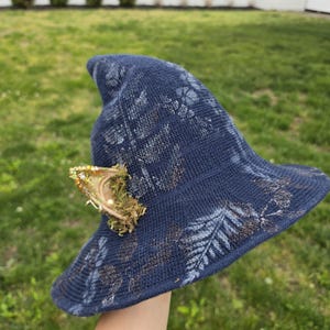 May include: A navy blue pointed hat with a textured knit surface featuring a light blue fern leaf pattern. A single decorative ear-shaped attachment is affixed to the side, adorned with green moss, gold glitter, and small white beads.