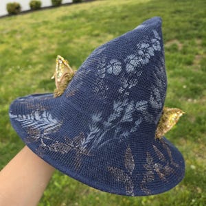 May include: A navy blue sun hat with a pointed crown and a wide brim. The hat is decorated with a floral pattern in white and gold accents. Two gold butterfly embellishments are attached to the brim.