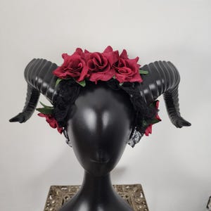May include: A black headband with two black horns and a red rose flower crown. The headband is on a black mannequin head.