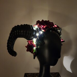 May include: A headpiece featuring large, curved, black ram horns and deep red artificial roses. The accessory is adorned with thin, glowing white LED fairy lights woven throughout the flowers and dark foliage, creating a luminous effect.